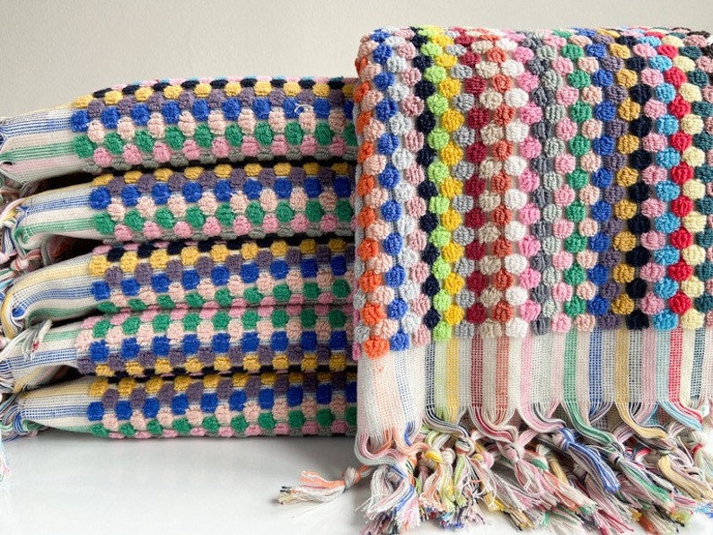 May include: A stack of colorful, textured towels. The towels feature a repeating pattern of raised dots in various colors, including blue, pink, yellow, and green. The edges have striped detailing and fringe.