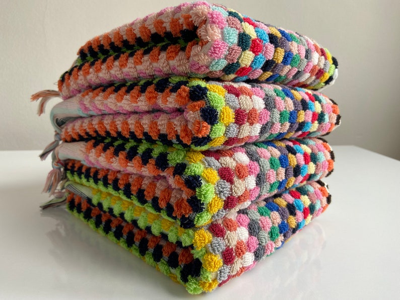 May include: A stack of four colorful, textured square towels. Each towel features a pattern of small, raised circles in various colors, including pink, orange, green, blue, and yellow. The towels have fringe on one side.