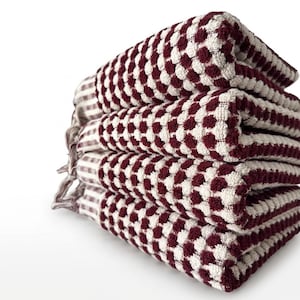 May include: A stack of four folded towels. The towels feature a textured pattern of maroon and white dots. The towels are neatly stacked, with the edges aligned. The background is white, creating a clean aesthetic.