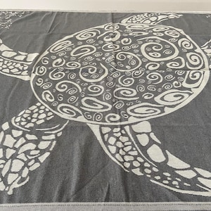 Peut inclure: Une serviette de plage grise avec un motif de tortue de mer blanche. La tortue a une carapace détaillée avec un motif en spirale.