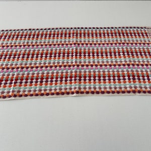 May include: A colorful kitchen towel with a checkered pattern and fringe edges. The towel features a mix of red, orange, pink, green, and white.