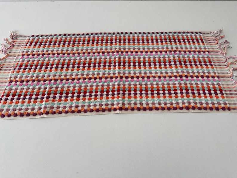 May include: A colorful kitchen towel with a checkered pattern and fringe edges. The towel features a mix of red, orange, pink, green, and white.