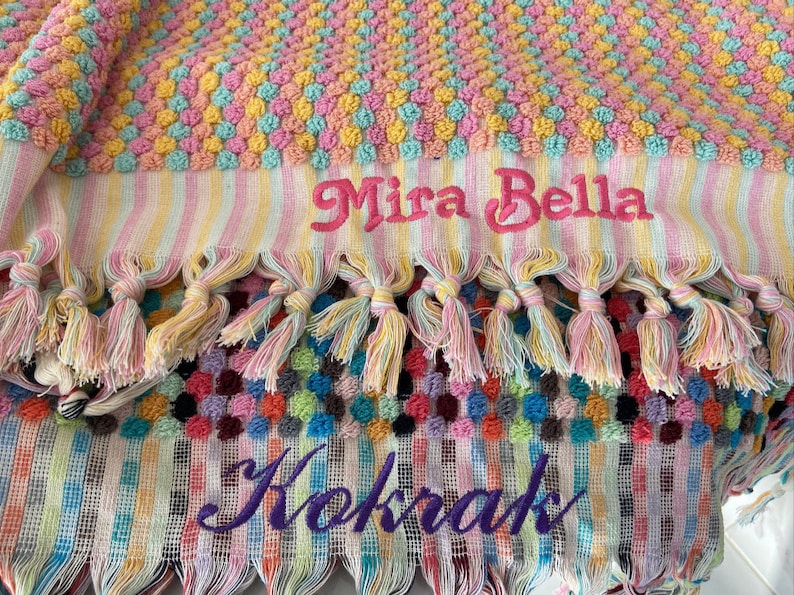 Pode incluir: Uma toalha de praia listrada colorida com um design de pompons rosa e amarelo. A toalha tem franjas e &eacute; bordada com os nomes "Mira Bella" e "Kokrak".