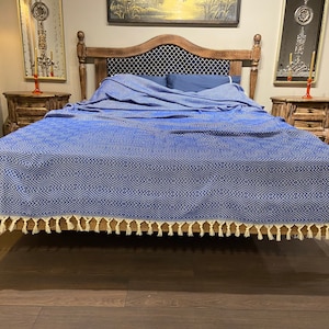 Może przedstawiać: Niebiesko-biały pled z frędzlami na drewnianym stelażu łóżka. Pled ma geometryczny wzór i jest wykonany z miękkiej, wygodnej tkaniny.