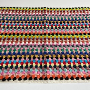 May include: A rectangular, multi-colored woven textile with a repeating pattern of raised, rounded squares. The color palette includes pink, orange, yellow, green, blue, and black, creating a vibrant and textured surface. The edges are finished with a white border.