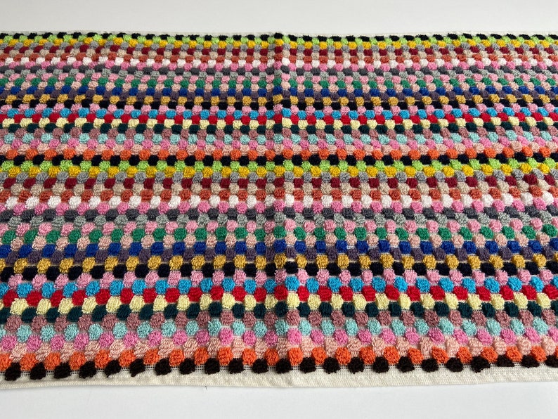May include: A rectangular, multi-colored woven textile with a repeating pattern of raised, rounded squares. The color palette includes pink, orange, yellow, green, blue, and black, creating a vibrant and textured surface. The edges are finished with a white border.