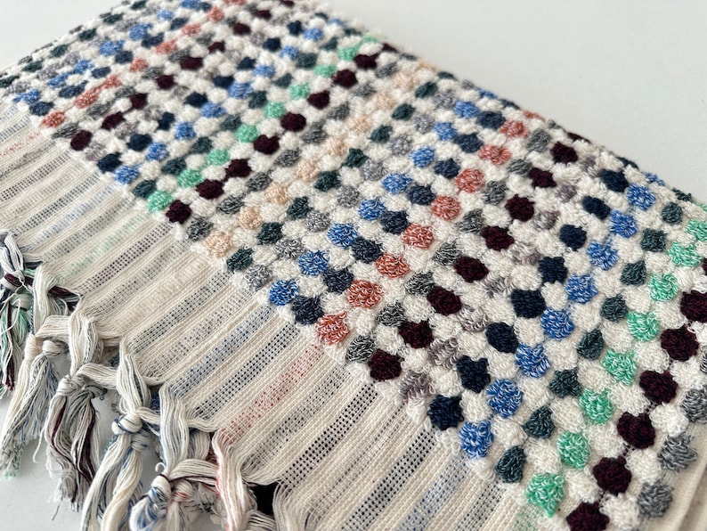 K&ouml;nnte beinhalten: Ein wei&szlig;es Badetuch mit einem farbenfrohen Muster. Das Muster besteht aus kleinen, erhabenen Kreisen in Blaut&ouml;nen, Gr&uuml;n, Rot und Braun. Das Handtuch hat Fransen an den Enden.