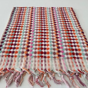 May include: A colorful patterned hand towel with a fringe border. The towel features a checkered pattern with small circles in shades of red, orange, green, pink, and purple.
