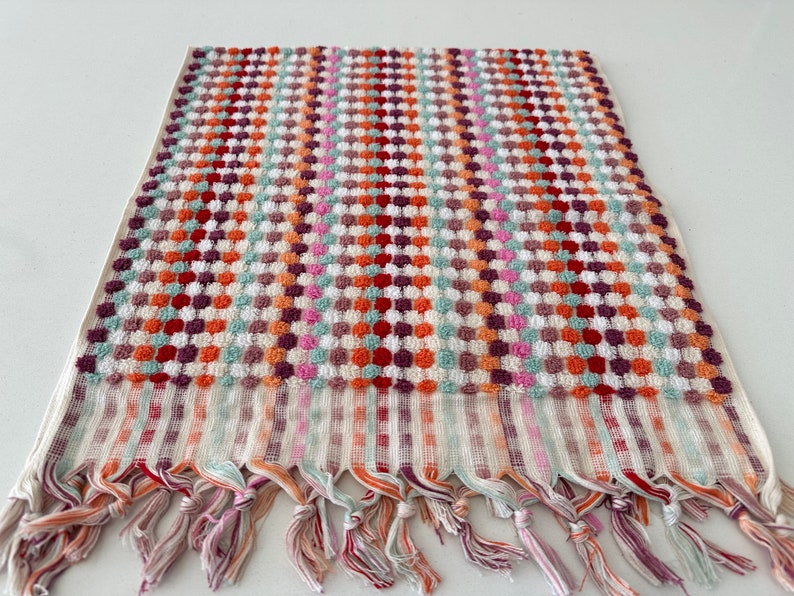 May include: A colorful patterned hand towel with a fringe border. The towel features a checkered pattern with small circles in shades of red, orange, green, pink, and purple.