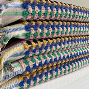 May include: A stack of four colorful woven towels. The towels feature a striped pattern with fringe on the ends. The top layer has a textured, dotted pattern in blue, green, pink, and yellow. The towels are folded neatly, showcasing the vibrant colors and textures.