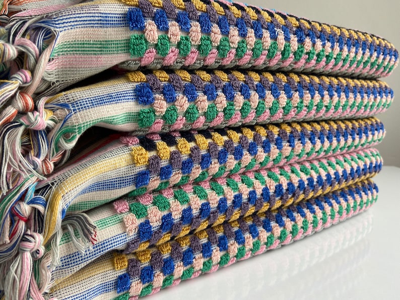 May include: A stack of four colorful woven towels. The towels feature a striped pattern with fringe on the ends. The top layer has a textured, dotted pattern in blue, green, pink, and yellow. The towels are folded neatly, showcasing the vibrant colors and textures.