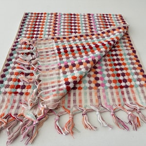 May include: A white bath towel with a colorful geometric pattern of red, orange, green, and purple. The towel has a fringe border and is folded in half.