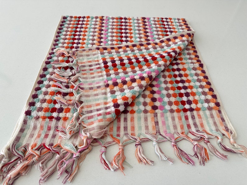 May include: A white bath towel with a colorful geometric pattern of red, orange, green, and purple. The towel has a fringe border and is folded in half.