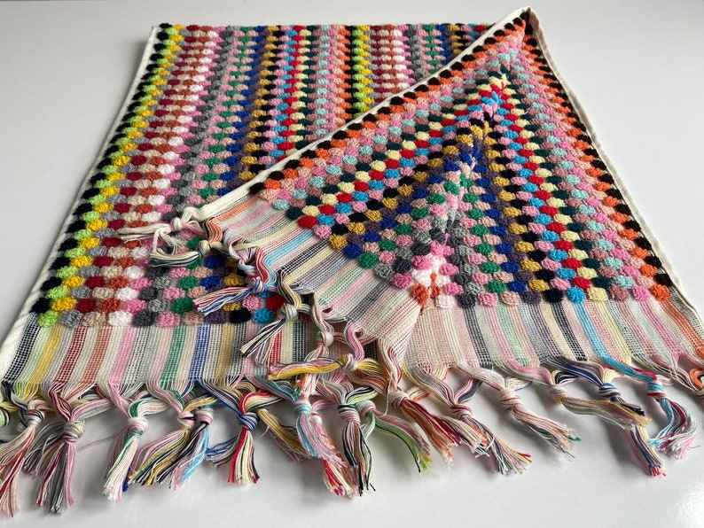 May include: A colorful hand towel with a multi-colored pom-pom design and fringe trim. The towel is folded in half and has a white background.