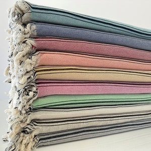 Könnte beinhalten: Ein Stapel von acht gestreiften Handtüchern in verschiedenen Farben, darunter hellblau, rosa, beige, grün und weiß. Die Handtücher haben einen Fransenrand und sind aus einem weichen, saugfähigen Material gefertigt.