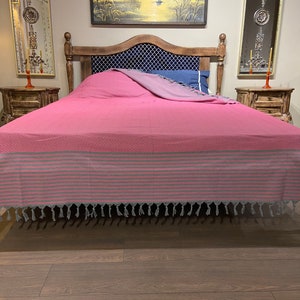 Puede incluir: Un cubrecama a rayas rosa y gris con borlas en un marco de cama de madera. El cubrecama tiene un patr&oacute;n de diamante y est&aacute; hecho de una tela suave y c&oacute;moda.