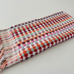 May include: A colorful hand towel with a checkered pattern and fringe. The towel is made of a soft, absorbent material and features a variety of colors, including red, orange, yellow, green, blue, and purple.