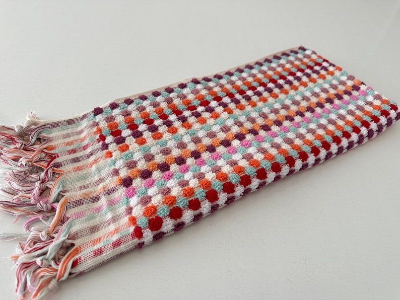 May include: A colorful hand towel with a checkered pattern and fringe. The towel is made of a soft, absorbent material and features a variety of colors, including red, orange, yellow, green, blue, and purple.