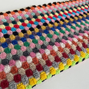 May include: A rectangular, multi-colored, textured fabric with a repeating pattern of raised, rounded squares. The color palette includes shades of pink, red, blue, green, yellow, and gray. The edges are finished with a contrasting border.