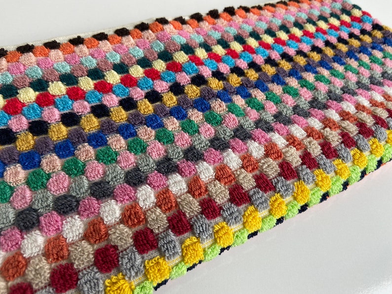 May include: A rectangular, multi-colored, textured fabric with a repeating pattern of raised, rounded squares. The color palette includes shades of pink, red, blue, green, yellow, and gray. The edges are finished with a contrasting border.