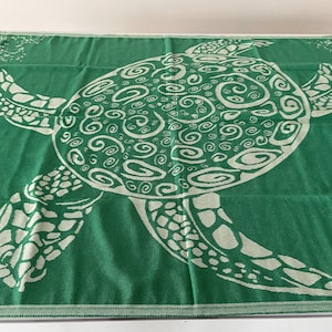 Peut inclure: Une serviette de plage verte avec un motif de tortue de mer blanche. La carapace de la tortue est décorée d'un motif en spirale.