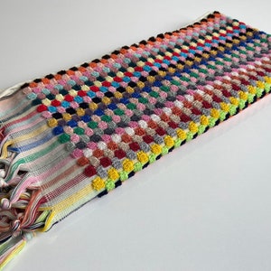 May include: A rectangular, multi-colored Turkish towel with a textured, raised dot pattern. The towel features a variety of colors, including pink, blue, yellow, and green. The ends have striped patterns and knotted fringe.