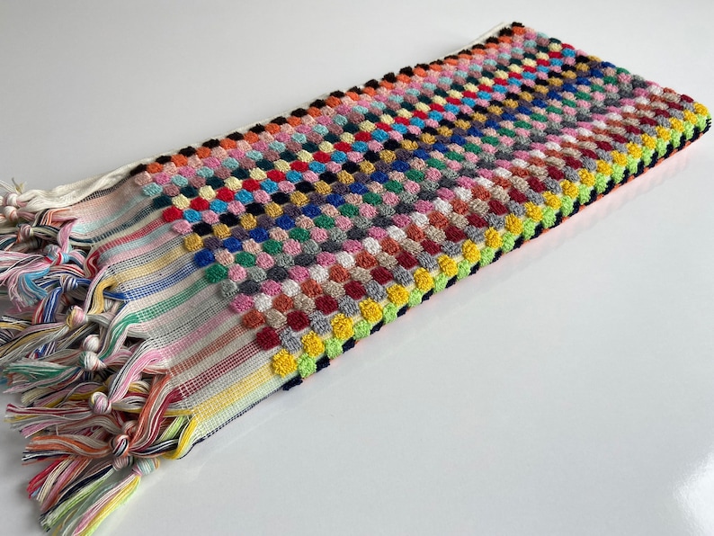 May include: A rectangular, multi-colored Turkish towel with a textured, raised dot pattern. The towel features a variety of colors, including pink, blue, yellow, and green. The ends have striped patterns and knotted fringe.
