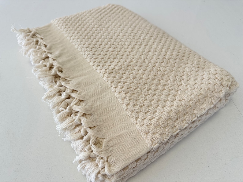 Pode incluir: Uma toalha de banho de tecido waffle de cor creme com uma borda de franjas. A toalha est&aacute; dobrada ao meio e tem uma superf&iacute;cie macia e texturizada.