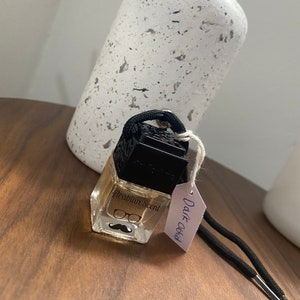 Puede incluir: Una pequeña botella de vidrio con una tapa negra y una cuerda negra. La botella contiene un líquido transparente y tiene una etiqueta que dice "Arabian Scent". La botella cuelga de una cuerda negra con una etiqueta blanca que dice "Dark Orchid".