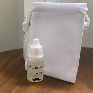 Puede incluir: Una pequeña botella de vidrio transparente con una tapa blanca y una etiqueta blanca que dice "Arabian Scent" con un gráfico de bigote y gafas negras. La botella está sobre una superficie de madera junto a una bolsa de cordón blanca.