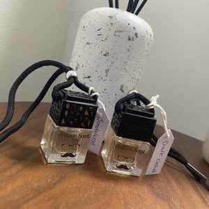 Puede incluir: Dos ambientadores para coche de cristal transparente con tapas negras y cordones negros. Los ambientadores tienen un gráfico de bigote y el texto "Arabian Scent". Una etiqueta dice "Sandalwood" y la otra "Dark Orchid".
