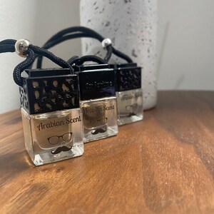Puede incluir: Tres ambientadores de coche de vidrio transparente y negro con una cuerda negra para colgar. Cada ambientador tiene un gráfico en blanco y negro de un bigote y gafas y el texto "Arabian Scent".
