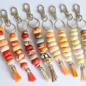 May include: A selection of keyrings with decorative beaded strands in various colours. Each keyring features a silver-tone clasp, a beaded strand with a tassel, and a ring. The beads are in shades of orange, red, yellow, and cream.
