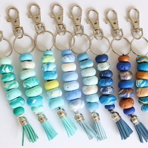 May include: A selection of handmade keyrings with blue and turquoise polymer clay beads. Each keyring has a silver-tone clasp, a metal ring, and a matching tassel. The beads feature marbled, speckled, and solid designs in various shades.