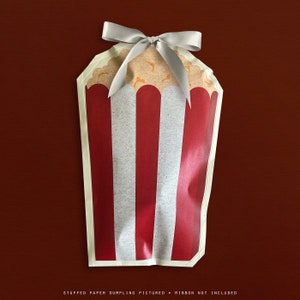 May include: A paper dumpling bag designed to look like a popcorn container, with red and white stripes. The top is filled with popcorn and tied with a silver ribbon. The text at the bottom reads: STUFFED PAPER DUMPLING PICTURED • RIBBON NOT INCLUDED.