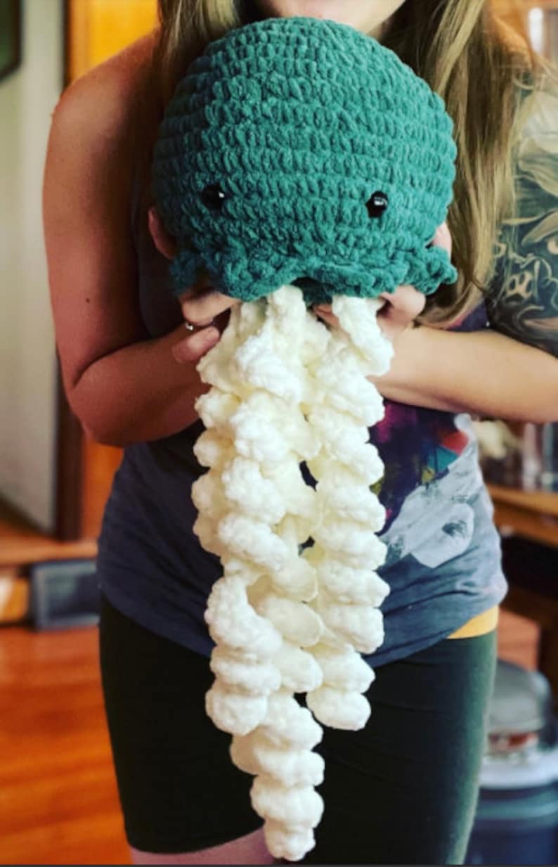 May include: A crocheted plush jellyfish toy with a teal-colored body and white tentacles. The jellyfish is being held by a person.