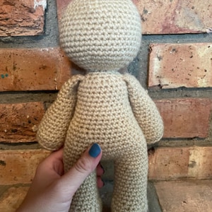 Peut inclure: Une poupée en crochet avec un corps et une tête beiges. La poupée est debout sur une surface en brique.