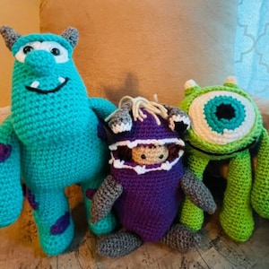 May include: Three crocheted plush toys inspired by the characters from the movie Monsters Inc. The toys are a blue monster with purple spots, a green monster with a large eye, and a purple monster with a white mouth.