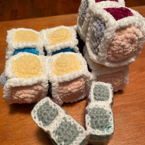May include: A crocheted toy made of multiple colorful cubes. The cubes are connected together and have a white border. The toy is made of soft, fuzzy yarn in shades of pink, yellow, blue, gray, and purple.