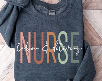 May include: A dark grey sweatshirt with the word "NURSE" printed in colourful letters. The text "labour & delivery" is written in white script below the word "NURSE".
