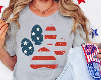 Może przedstawiać: Szary T-shirt z nadrukiem odcisku łapy w stylu amerykańskiej flagi. Modelka nosi niebieskie dżinsy i trzyma parę czerwono-biało-niebieskich okularów przeciwsłonecznych.