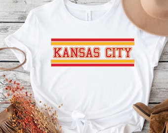 Puede incluir: Camiseta blanca con rayas rojas y doradas y el texto "KANSAS CITY" en negrita.