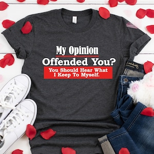 May include: A dark gray t-shirt with the text "My Opinion Offended You? You Should Hear What I Keep To Myself." printed in white. The shirt is laid out on a white surface with red rose petals scattered around it.
