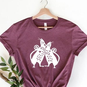 Może przedstawiać: Bordowy t-shirt z białym nadrukiem przedstawiającym dwie szczury, które się obejmują. Szczury mają serca na klatce piersiowej.