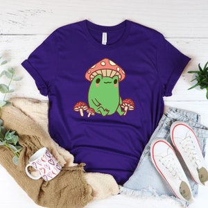 May include: Purple t-shirt with a cartoon frog wearing a red and white mushroom hat. The frog is sitting on a mushroom and surrounded by other mushrooms.