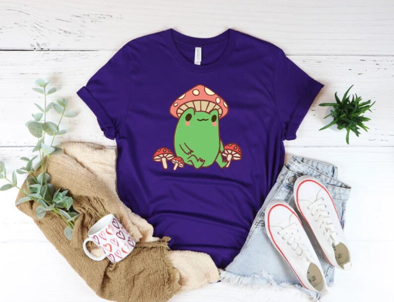 May include: Purple t-shirt with a cartoon frog wearing a red and white mushroom hat. The frog is sitting on a mushroom and surrounded by other mushrooms.