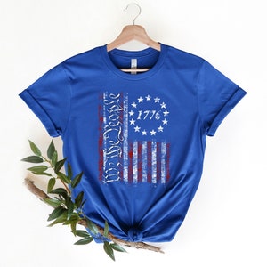 K&ouml;nnte beinhalten: Ein k&ouml;nigsblaues T-Shirt mit einem abgenutzten Design einer amerikanischen Flagge. Die Flagge hat rote und wei&szlig;e Streifen und einen Kreis aus Sternen mit dem aufgedruckten Text "We the People" und "1776".