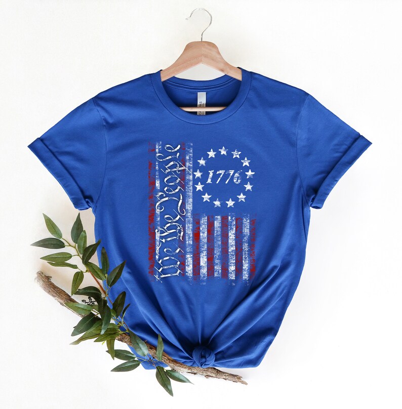 K&ouml;nnte beinhalten: Ein k&ouml;nigsblaues T-Shirt mit einem abgenutzten Design einer amerikanischen Flagge. Die Flagge hat rote und wei&szlig;e Streifen und einen Kreis aus Sternen mit dem aufgedruckten Text "We the People" und "1776".