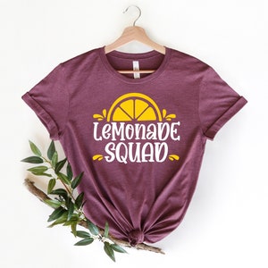 May include: A maroon t-shirt with a yellow lemon graphic and the text "Lemonade Squad" in white.