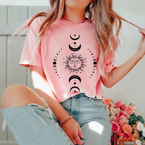 May include: A pink t-shirt featuring a black sun and moon design. The sun has a face and the moon is depicted in various phases.